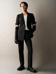 A person stands confidently against a neutral backdrop, wearing a dark suit with rolled-up sleeves, showcasing a white inner shirt. Black gloves and chunky shoes complete the ensemble. The individual exudes a stylish and modern vibe, blending classic and contemporary fashion elements, with a relaxed yet poised stance, evoking a sense of effortless elegance and sophistication.