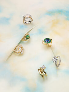 A collection of six rings is elegantly arranged on a soft, pastel background with swirling blue and beige hues. The rings feature intricate designs, showcasing a variety of gemstones, including vibrant green and deep blue stones, along with clear, sparkling diamonds. The bands vary in style, some gold, others silver, creating a luxurious and artistic display.