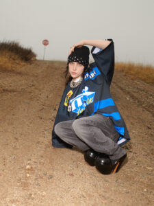 A person crouches on a gravel path, wearing a black beanie with studs, a chain necklace, oversized jersey, and gray jeans. Their hand rests on their beanie. The background features dry grass and a misty sky with a red octagonal sign in the distance. The setting has a rugged, natural feel, contrasting with the person's bold attire.