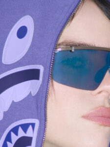 A person is wearing reflective blue sunglasses and a purple hoodie zipped up partially over the face. The hoodie features a stylized design resembling a shark with eyes and teeth. The close-up view highlights the person's eye through the glasses and their lips, suggesting a fashion-forward, edgy style with attention to detail and color contrast.