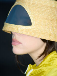 A person is wearing a unique woven headpiece that covers the upper face, featuring a transparent visor. The headpiece resembles a futuristic visor blending style and functionality. The clothing is yellow, matching the headpiece's tone, creating a cohesive look. Soft lighting highlights the facial features, while the background is a deep black, adding contrast to the ensemble.