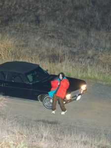 A person stands beside a black vintage car on a dirt path, surrounded by dry grass and rugged terrain. They wear a bright orange and blue jacket with brown pants and beige shoes. It's nighttime, with the car's headlights illuminating the scene. The atmosphere feels quiet and slightly mysterious, with a sense of adventure.