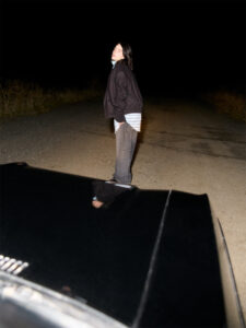 A person stands on a gravel road at night, illuminated by headlights, with tall grass on either side. Dressed in a casual jacket and striped shirt, they lean slightly, gazing upward. The foreground features the reflective surface of a car hood, capturing part of their reflection. The atmosphere is quiet and isolated, suggesting a moment of contemplation.