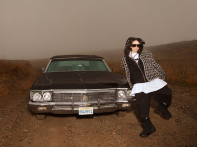 A person leans against a classic black car parked on a dirt path. The background features a foggy, overcast sky, and dry grass stretches across the landscape. The individual is dressed in layered, casual attire, with a houndstooth-patterned coat, black hoodie, and oversized sunglasses, exuding a relaxed, stylish vibe.