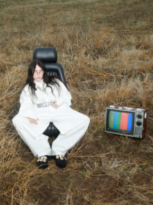 In a grassy, dried-out field, a person with long dark hair reclines in a detached car seat, dressed in a white suit with ornate jewelry and embellished black shoes. Beside them, an old television set displays colorful vertical stripes on the screen, contrasting against the earthy tones of the surroundings. The scene evokes a surreal, contemplative atmosphere.