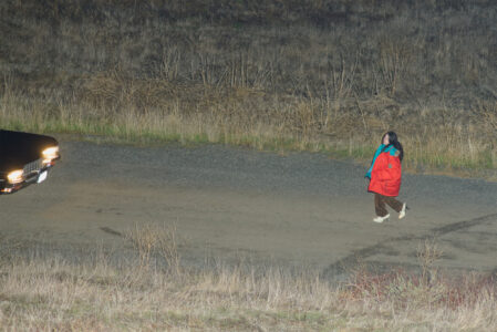 A person walks on a gravel path, wearing a large red and teal jacket with dark pants and white shoes. The background features dry grass and shrubs on a sloping terrain. Approaching from the left, the headlights of a dark vehicle illuminate the scene, casting a glow amidst the quiet, open landscape at night.
