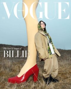 A person in a hooded jacket and glasses stands in a field, wearing oversized pants and facing upwards. They lean against a gigantic sculpture of a woman’s leg in a red high heel, with dry grass stretching into the distance. The sky is clear, creating a juxtaposition of scale and fashion in the open landscape.