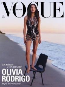 A young woman stands barefoot on a simple chair placed on a quiet beach at dusk. The ocean stretches beside her under a soft gradient sky. She wears a striking black outfit adorned with reflective, sculptural embellishments that catch the fading light. Her posture is steady and confident, gaze forward, creating a bold contrast between natural calm and high-fashion intensity.