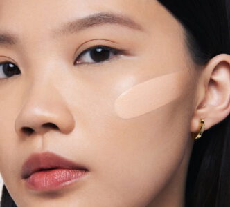 A close-up view of a person's face showcases smooth, clear skin with a neutral expression. The complexion appears even-toned with a makeup swatch applied on the cheek, highlighting a subtle glow. The person has dark eyes with natural lashes and well-groomed eyebrows. Dark, straight hair frames the face, and a small hoop earring adorns the ear.