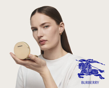 A person with smooth, dark hair parted in the center holds a round, gold compact mirror delicately in an open palm. They wear a simple white shirt and small gold hoop earrings. Their expression is serene and poised against a soft, neutral background, exuding elegance and sophistication with a focus on beauty and grooming.