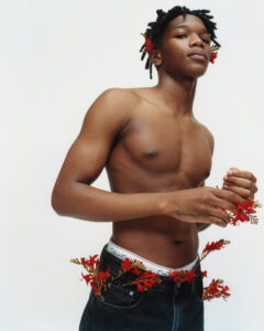An individual with a confident expression stands against a plain backdrop. Their hair is styled in twists adorned with red flowers. They're shirtless, with floral embellishments tucked into the waistband of their dark jeans. A tattoo is visible on their wrist. The overall vibe is a fusion of natural elements with modern style, exuding confidence and creativity.