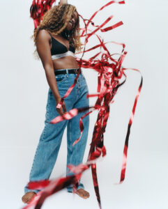 A person with long curly hair is wearing a black bralette and loose-fitting blue jeans. They are standing on a light background, surrounded by flowing red and black ribbons. The ribbons create a dynamic and lively effect, adding a sense of movement and celebration to the scene. The overall composition is vibrant and energetic.