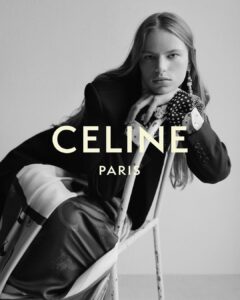 A person with long hair poses in a black and white setting, leaning on a rustic chair. They wear a tailored jacket and a patterned blouse with gloves featuring polka dots and floral designs. The attire includes a flowing skirt with bold graphics. The overall composition is elegant and artistic, emphasizing modern fashion in a minimalist environment.