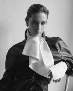 A person exudes elegance with a polished, slicked-back hairstyle, wearing a bold leather outfit accented by a large, dramatic white scarf. The pose is poised and confident, juxtaposed against a textured wall, creating a stark contrast. The monochrome tone enhances the sophisticated and artistic aura, emphasizing the modern, minimalist style and intense gaze.
