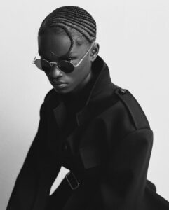 A person with tight cornrows and sunglasses gazes downward. The monochrome setting amplifies the intensity and focus of their expression. Wearing a structured coat with a high collar, they exude sophistication and confidence. The lighting accentuates the textures of their hairstyle, coat, and accessories, creating a striking, modern, and stylish image with a contemporary and artistic vibe.