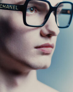 A close-up of a person's face features large, rectangular eyeglasses with a glossy finish. The gaze is directed forward, with eyebrows subtly arched. The complexion is smooth, with softly hued lips. The lighting casts a gentle glow, highlighting the details of the glass frames and focusing on the quiet intensity in the eyes.