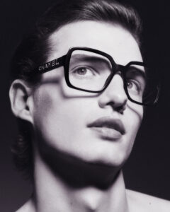 A person with a focused expression gazes upward. They have short, styled hair, and are wearing large, square-framed glasses with a distinctive design. The lighting highlights their facial features, casting soft shadows. The monochromatic tones add elegance and sophistication to the scene, creating a classic and timeless feel. The overall mood is contemplative and stylish.