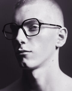 A person with a light complexion is depicted with closely cropped hair and large black-rimmed glasses. The lighting casts sharp shadows, creating a dramatic and artistic contrast. The overall tone is monochromatic, highlighting the structural elements of the glasses. The person gazes slightly away, capturing a sense of introspection and modern minimalism.