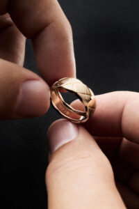 Two fingers delicately hold a polished gold ring, which features a quilted pattern on its surface. The background is a soft black, creating a contrast that emphasizes the ring's shine. Light highlights its smooth curves and intricate design. The ring's inner band shows craftsmanship details, reflecting elegance and care. Each element suggests timeless sophistication and precision.