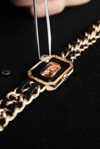 A gold watch with a transparent caseback is being closely inspected. The intricate inner mechanisms are visible, showcasing delicate gears and components. A pair of metal tweezers is holding a small circular piece above the watch. The watch's strap features a bold design with intertwined gold and black links, contrasting with the dark textured surface beneath it.