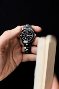 A hand is elegantly holding a sleek, black watch with a shiny metallic band and clear, bold numerals on its face. The watch is displayed against a dark background, emphasizing its polished design. Adjacent to the hand, pages of an open book are visible, suggesting a moment of casual sophistication and leisure.