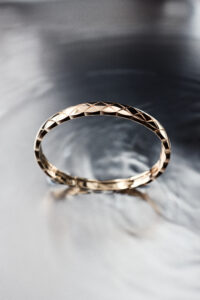 A delicate golden ring with a faceted pattern reflects a soft light, creating subtle highlights and shadows. The ring appears to be elegantly crafted, with attention to detail in its geometric design. It rests on a smooth, reflective surface that captures gentle ripples of light, adding an ethereal quality to the scene, emphasizing the ring's sophisticated allure.