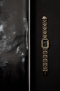 A luxurious wristwatch is elegantly displayed against a dark, textured surface. Its face is black, accented with a gold frame, which complements the intertwined black and gold links of the strap, forming a sophisticated luxury design. To the left, an area of water or liquid adds a touch of contrast with subtle reflections and ripples.