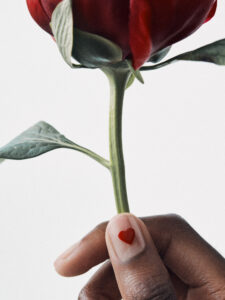A hand gently holds a single red rose by its slender green stem. The rosebud is vibrant and fresh, surrounded by a few lush leaves. The person's fingernail is adorned with a small red heart, symbolizing love and attention to detail. The background is a simple, soft white, highlighting the elements beautifully.