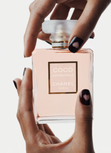 A pair of hands gracefully holds a rectangular glass bottle containing a delicate peach-colored liquid. The bottle has a square, translucent cap, and its glass structure exudes elegance. The fingernails are painted dark, each featuring a heart-shaped cutout, adding a touch of playful sophistication. The composition highlights luxury and style.