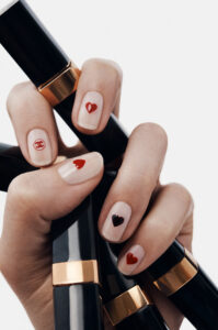 A hand with manicured nails holds several black and gold-capped cylindrical objects. The nails are adorned with small red and black hearts on a natural, nude base. The overall composition highlights elegance and attention to detail, with the contrast between the shiny cylinders and the delicate artwork on the fingernails, creating a sophisticated and chic aesthetic.