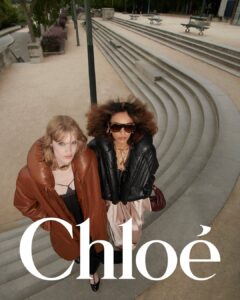 Two individuals stand confidently in a stylish setting, wearing oversized puffer jackets. One sports a brown jacket with blonde hair, while the other has a black jacket with voluminous, curly hair and sunglasses. They are surrounded by a modern, outdoor environment featuring curved concrete steps and symmetrical design elements, creating a striking contrast with their fashionable attire.