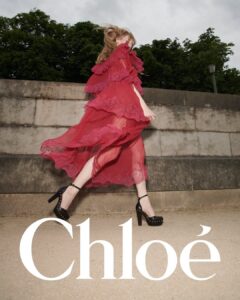 A person is moving energetically, wearing a flowing red dress with lace details. The dress billows around them, revealing black high-heeled shoes with ankle straps and studs. The background features a stone wall and lush green trees under a cloudy sky. The dynamic pose and vibrant attire create a sense of motion and elegance.