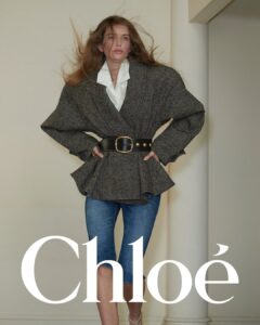 A person stands with a confident pose, hands on hips. They wear a gray, textured coat with a broad belt, emphasizing the cinched waist. The coat's wide shoulders and peplum hem accentuate the silhouette. Underneath, a white collared shirt is visible. Blue, knee-length denim pants complete the look. Wind blows their long hair, adding dynamic movement.