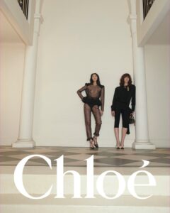 Two individuals stand elegantly in a minimalistic, high-ceilinged room with pillars. One wears a sheer, polka-dotted ensemble, while the other dons a black outfit with capri pants, holding a handbag. The muted color palette, patterned floor, and architectural elements create a sophisticated and stylish ambiance. Both exhibit confident postures, reflecting a blend of modernity and elegance.