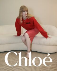 A person with long, blond hair sits on a pale, textured couch, wearing a vibrant red sweater and matching red skirt. They have an intense, focused gaze directed to their left. Gold jewelry, including a necklace and bracelet, accents their outfit. The neutral-toned background and patterned carpet create a stylish, contemporary setting.