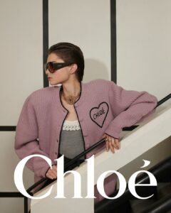 A person with slicked-back hair stands confidently, wearing oversized sunglasses and a chunky necklace. They are dressed in a pink, textured cardigan over a gray top, leaning against a white railing with black accents. The pose and setting suggest a modern, stylish ambiance, blending fashion-forward elements with a classic, neutral backdrop for a striking effect.