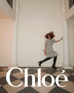A stylish woman with voluminous curly hair strikes a dynamic pose between two white columns. She's wearing a black-and-white polka dot dress paired with knee-high black boots. The floor beneath her features a checkerboard pattern of light and dark tiles. The setting is minimalist, with a soft, neutral wall as the backdrop.