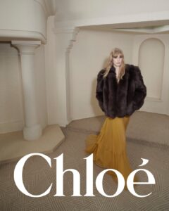 A blonde woman stands in an elegantly designed room with cream walls and a patterned carpet. She wears a voluminous dark fur coat and a flowing yellow skirt that cascades to the floor. Architectural elements include a column and arch, adding sophistication to the setting. Her expression is poised, contributing to the graceful and refined atmosphere.