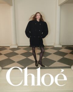 A person stands confidently, clad in an oversized black coat with button detailing. Their long hair flows naturally over their shoulders. Black tights and elegant heels complete the ensemble. The setting features a classic interior with white columns and a checkered floor pattern. The atmosphere is sophisticated and stylish, capturing a sense of modern elegance.