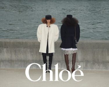 Two individuals stand by a stone wall next to water. One wears a white coat, dark trousers, black boots, and a black hat, with their hands in pockets. The other person, seen from behind, wears a dark coat, lace dress, brown boots, and a black hat. Both have voluminous hairstyles, with the backdrop of a river.