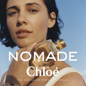 A person with short dark hair gazes into the distance against a clear blue sky. They hold a bottle with a rounded design, featuring golden accents and intricate details. The lighting highlights their natural complexion and calm expression. The setting suggests a warm environment, reflecting tranquility and elegance, while the object they hold draws focus and interest.