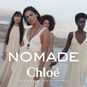 Four women pose gracefully by the water with a soft, serene backdrop. They wear flowing, light-colored dresses, exuding elegance and a sense of unity. The diverse group radiates confidence and natural beauty, standing against a landscape with blurred hints of mountains and sky. Their expressions are calm, complementing the tranquil and dreamy setting overall.