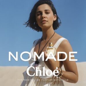 A woman stands against a cloudless blue sky, wearing a flowing white dress. Her hair is short and dark, softly blowing in the wind. She holds a bottle with a distinctive design. Her expression is serene and confident. The setting radiates warmth, suggesting a desert-like landscape with softly rolling dunes in the background.