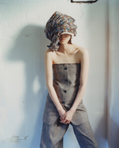 A person stands against a light-colored brick wall, wearing a strapless plaid outfit featuring buttons down the front. A large, floral fabric covers their head, obscuring the face, creating a dramatic silhouette. The hands are gently clasped in front, conveying a sense of calm and poise. Soft lighting casts subtle shadows, adding depth to the scene.