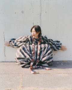A person is crouched against a cracked wall, wearing a voluminous plaid dress with long sleeves and a wide skirt. Sunlight casts soft shadows, highlighting the fabric's flowing texture and intricate patterns. Their hands touch the wall for balance, and the overall mood feels expressive and contemplative, capturing a blend of elegance and introspection.