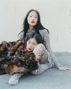 Two individuals are posing, showcasing fashion with a bold, feline-inspired pattern on their outfits. One sits on the ground, partially wrapped in an animal print faux fur coat. The other stands slightly behind, wearing a matching outfit. Both display an expression of calm elegance with dark hair and striking makeup, set against a plain, light backdrop.