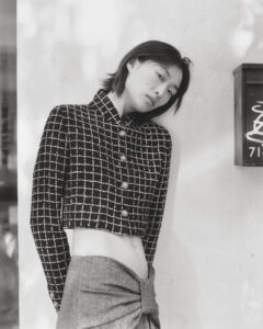 A person is leaning against a light-colored wall, dressed in a stylish ensemble. The outfit features a patterned, button-up jacket with a checkered design, and a high-waisted skirt tied in a knot. The individual has shoulder-length hair and a relaxed posture. The lighting casts soft shadows, creating a calm, elegant atmosphere. A rectangular box is nearby.
