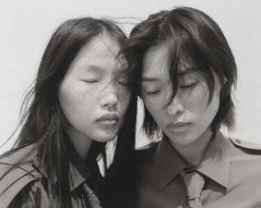 Two individuals share an intimate moment with their eyes closed, heads gently resting together. Their serene expressions suggest a deep connection. Both wear similar button-up shirts, and their flowing hair cascades naturally. The backdrop is bright, highlighting the calm ambiance. The monochromatic tones emphasize a sense of timelessness and tranquility in their poised, contemplative posture.