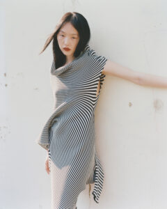 A person stands against a light-colored wall, wearing a black and white striped outfit with an asymmetrical design. The fabric drapes elegantly, accentuating the figure's posture. Long, dark hair flows freely, and a contemplative expression is visible. Soft light casts gentle shadows, creating a serene, minimalist atmosphere around the subject. There's a sense of calm and poise.