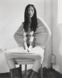 A monochrome scene features a person seated on a chair with a contemplative expression. They wear a draped garment, enveloping their arms. Long, dark hair frames their face. The setting suggests a minimalist, possibly artistic space, with light streaming through a window, enhancing the serene yet introspective mood. The floor has a rustic, distressed appearance.
